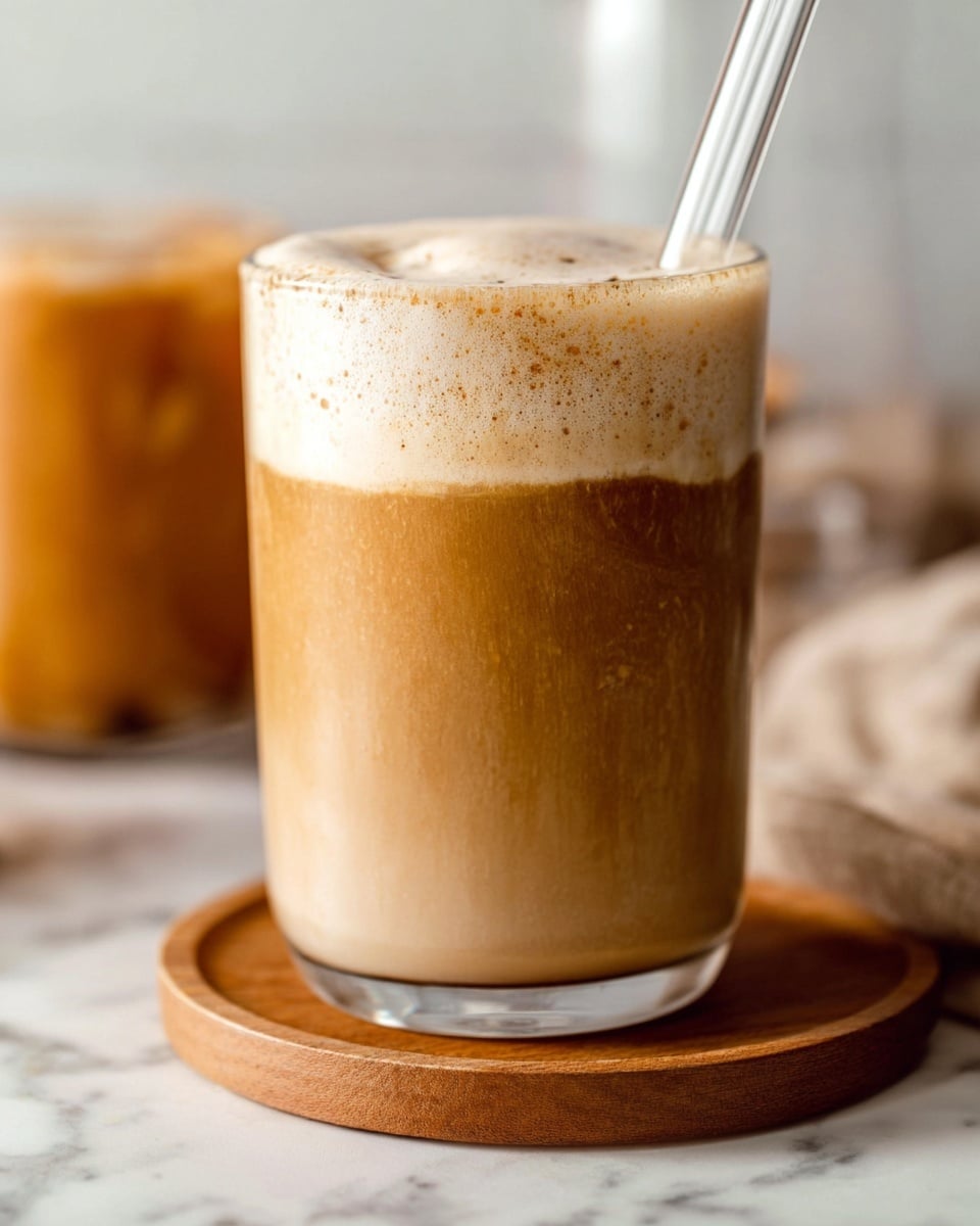 The image shows a clear glass filled with a creamy layered drink. The bottom layer is a smooth, light brown liquid, making up most of the glass. On top, there is a thick, foamy layer that is a lighter cream color with tiny brown specks, adding texture. A clear straw is placed inside the drink on the right side. The glass sits on a round wooden coaster, and a blurred second glass with the same drink is visible in the background. The whole scene is set against a white marbled surface. photo taken with an iphone --ar 4:5 --v 7