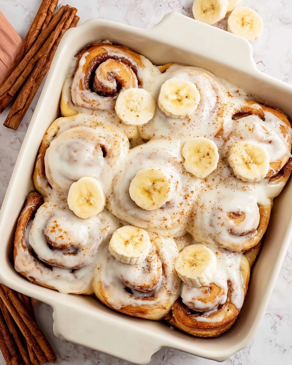 A white square baking dish holds ten cinnamon rolls covered in a thick layer of smooth, creamy white icing. The cinnamon rolls show swirls of golden brown dough visible beneath the icing with some darker cinnamon lines peeking through. Several banana slices are placed on top, their pale yellow color contrasting with the frosting. A light dusting of cinnamon powder is sprinkled over the rolls and banana slices. The dish sits on a white marbled surface with sticks of cinnamon nearby. Photo taken with an iphone --ar 4:5 --v 7
