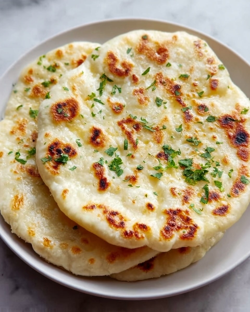 Two round flatbreads stacked on a white plate with a slightly crispy, golden-brown spotted surface. The flatbreads have a soft, fluffy texture with some light puffiness and small bubbles scattered throughout. Fresh small green herbs are sprinkled on top, adding a touch of color contrast to the creamy off-white dough. The plate is set on a white marbled surface. photo taken with an iphone --ar 4:5 --v 7