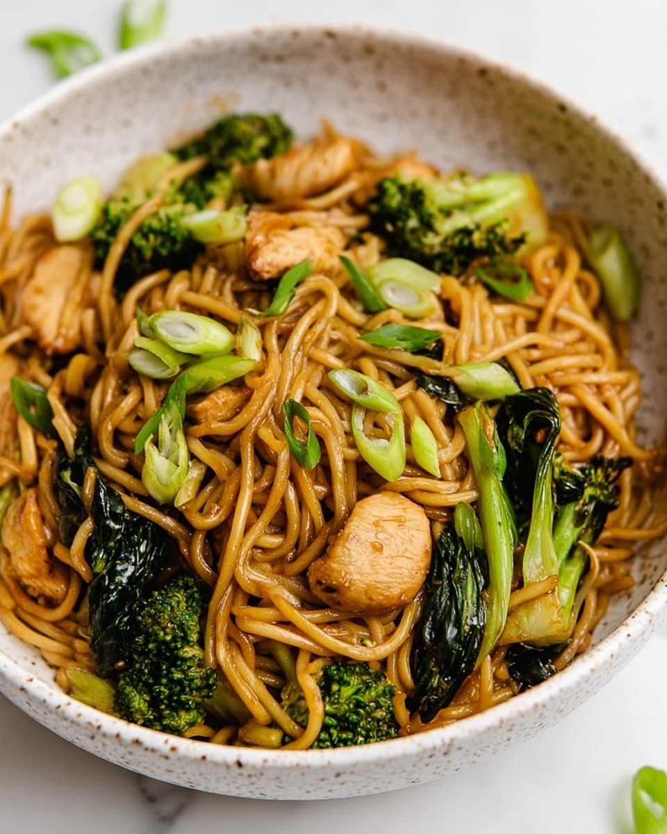 A white speckled bowl filled with stir-fried noodles that have a light brown, glossy coating, mixed with bright green broccoli florets and dark green bok choy stalks scattered throughout. There are chunks of light golden brown chicken pieces nestled among the noodles and vegetables. Thin slices of green onion are sprinkled on top, adding freshness and contrast to the dish. The bowl sits on a white marbled surface with a few green leaves blurred in the background, making the warm colors of the food stand out. Photo taken with an iphone --ar 4:5 --v 7