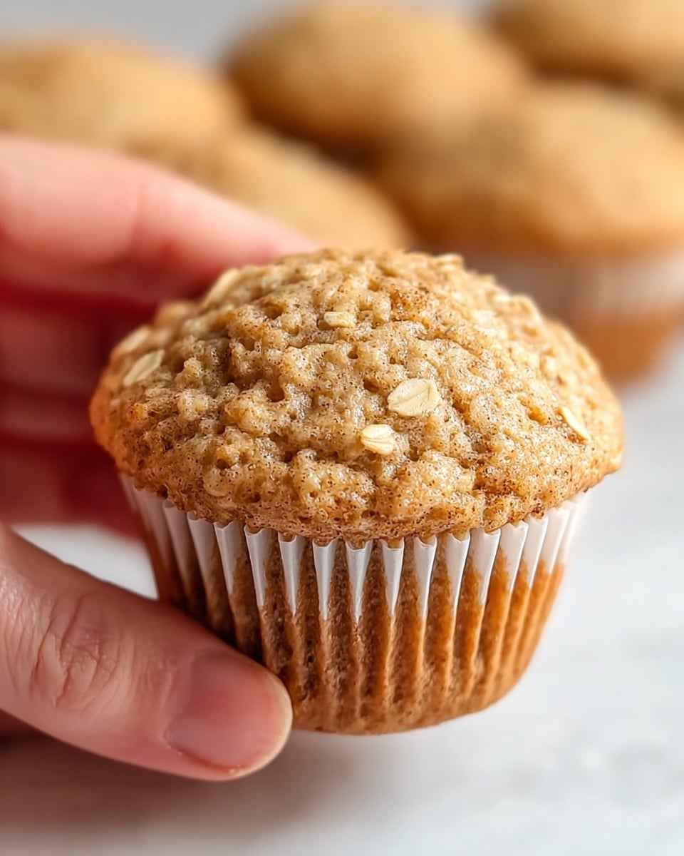A close-up image of a single oatmeal muffin held by a woman's hand, showing its slightly domed top with visible oat flakes embedded in the light golden-brown textured surface. The muffin has a soft, airy crumb with a few tiny specks of cinnamon or spice throughout. It sits inside a white paper muffin liner with distinct ridges wrapping around the base. The blurred background shows more similar muffins, all on a white marbled surface. The lighting is soft and natural, highlighting the texture and color of the muffin. photo taken with an iphone --ar 4:5 --v 7