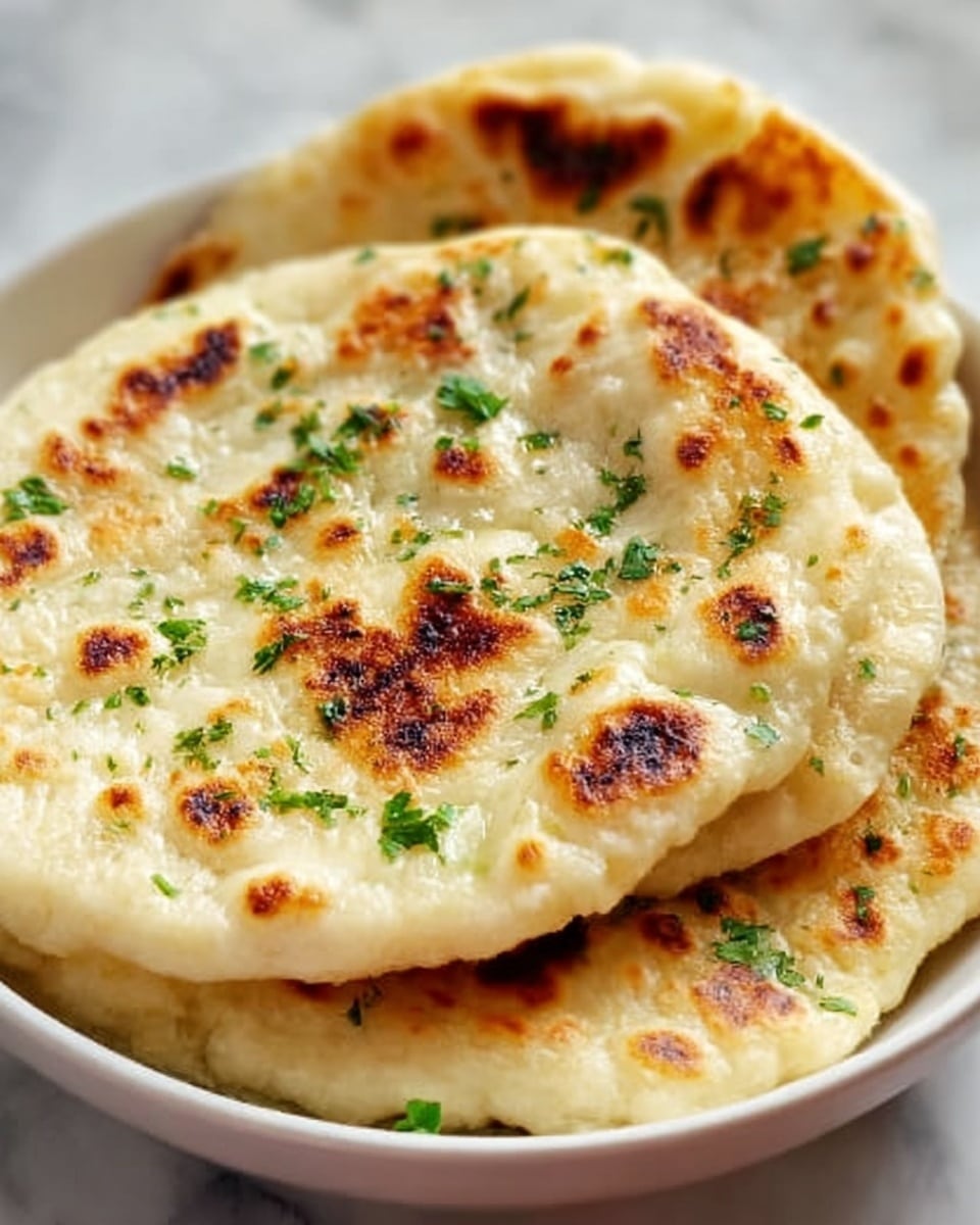 The image shows two round flatbreads stacked in a white bowl on a white marbled surface. The flatbreads have a light golden color with some darker brown spots from cooking. They look soft and slightly puffy, sprinkled with small green herb pieces on top, which add a fresh touch. The edges are slightly uneven, showing the handmade texture. The photo is close-up, highlighting the bread's toasted surface and soft layers inside. photo taken with an iphone --ar 4:5 --v 7