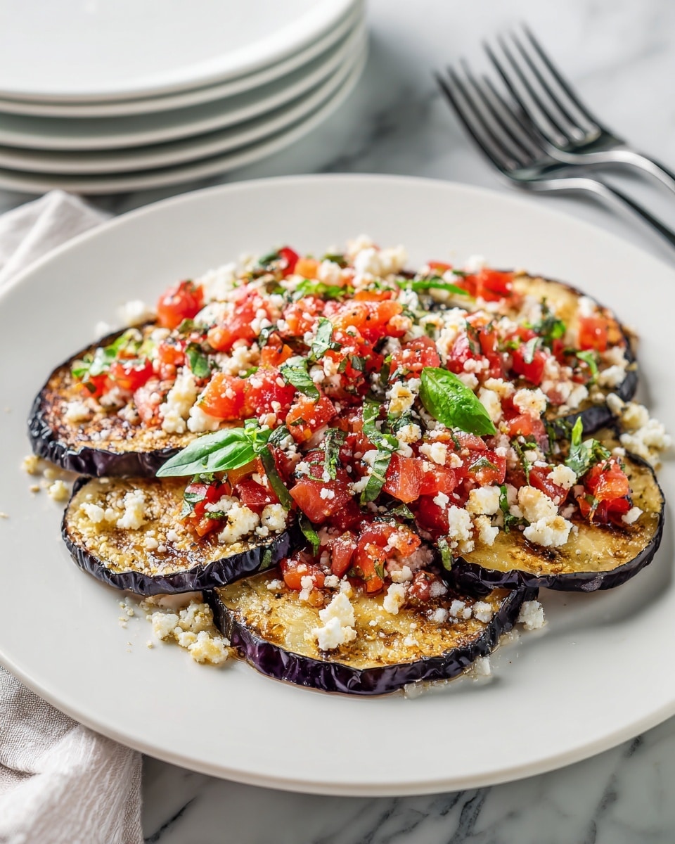 The image shows a white plate with several thin grilled eggplant slices arranged in a slight overlapping layer. The eggplant slices have a light brown, slightly crispy texture with dark purple edges. On top, there is a generous layer of small white crumbles of cheese, mixed with finely chopped bright red tomatoes. Fresh green basil leaves are scattered over the dish, adding a pop of color and freshness. The plate is placed on a white marbled surface, with stacked white plates and a fork visible in the background. Photo taken with an iphone --ar 4:5 --v 7