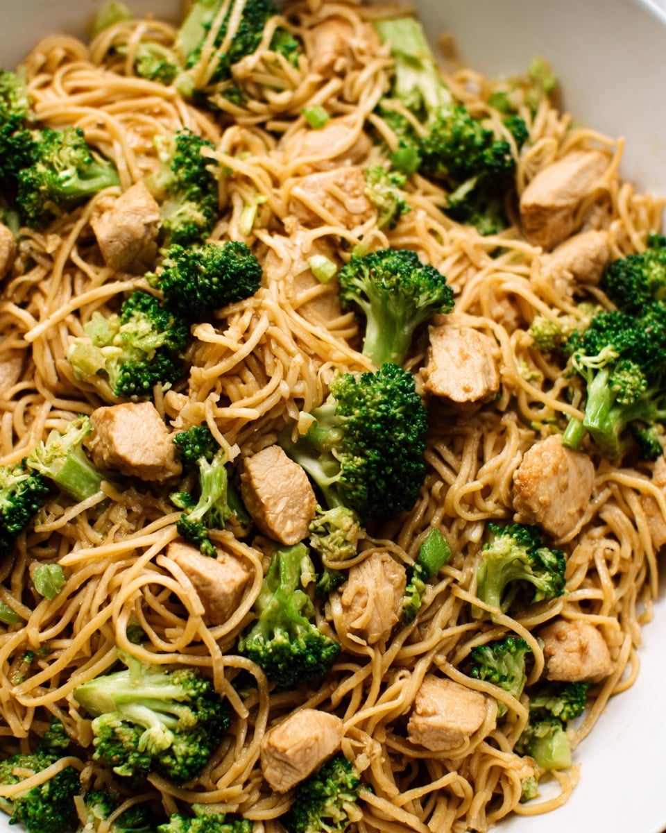A close-up image of cooked noodles mixed with green broccoli florets and light brown pieces of chicken, all layered evenly throughout the dish. The noodles are soft and slightly shiny with a few green broccoli pieces scattered on top and within the noodles. The chicken pieces are small, round, and slightly browned, placed evenly across the noodles. The dish is served on a white plate against a white marbled background. Photo taken with an iphone --ar 4:5 --v 7
