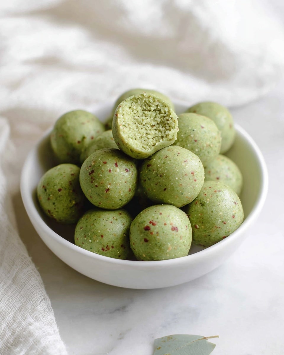 A woman's hand is holding two small round green balls with a smooth surface, one whole and one with a bite taken out. The bitten ball shows a crumbly, textured inside with a light green color and some small darker green and brown flecks. The balls rest above a blurred white plate in the background with more of the green balls inside, placed on a white marbled surface with a soft-focus white cloth. Photo taken with an iphone --ar 4:5 --v 7