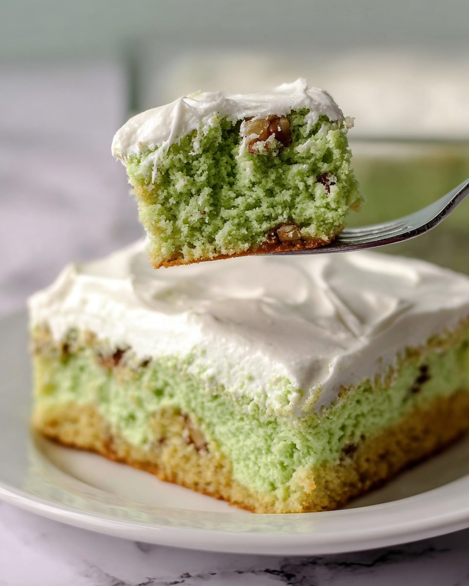 A close-up of a square slice of soft green cake with small brown nut pieces inside, topped with a thick layer of smooth white frosting. The cake layer is moist and crumbly, while the frosting looks creamy and fluffy. A small piece of the cake is lifted on a silver fork, showing the dense texture and the sharp contrast between the pale green cake and the white frosting. The slice sits on a white plate with a white marbled texture background. photo taken with an iphone --ar 4:5 --v 7