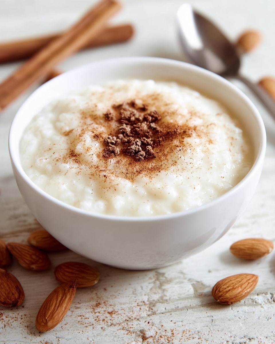 A white bowl with blue patterns is filled with thick, creamy rice pudding topped with a light dusting of brown cinnamon powder. The rice grains are soft and mixed well in the smooth pudding, creating a textured surface. A spoon is placed in the bowl, resting on the right side, with a woman's hand holding it gently. The bowl sits on a white marbled surface. photo taken with an iphone --ar 4:5 --v 7