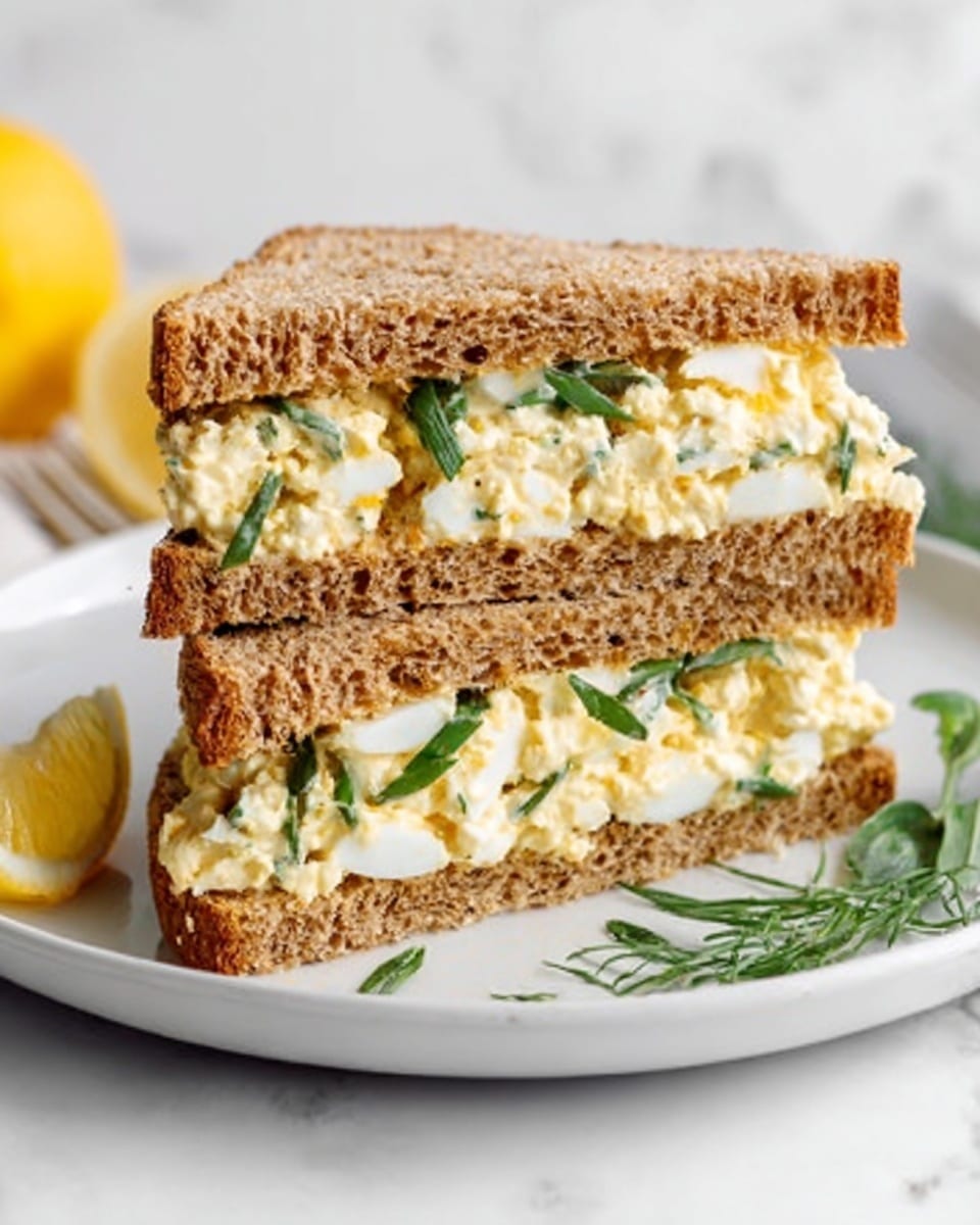 The image shows two sandwich halves placed side by side on a white plate. Each sandwich has three layers of brown whole grain bread, with a creamy egg salad filling visible between the slices. The egg salad is light yellow with small bits of green herbs mixed in, and the texture looks soft and creamy. On the plate near the sandwiches, there is a small wedge of lemon and some green herb sprigs for garnish. The background is a white marble surface. Photo taken with an iphone --ar 4:5 --v 7