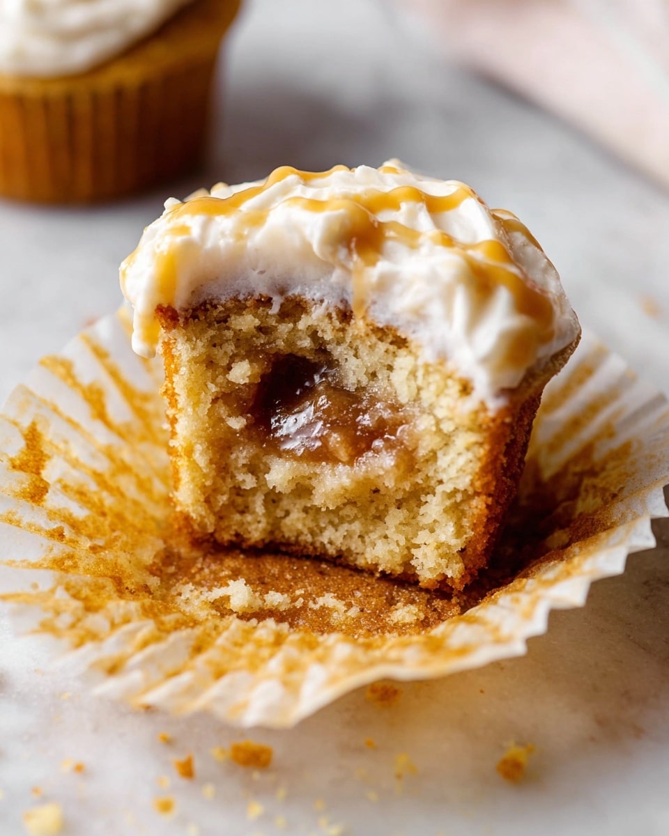 A close-up of a half-eaten cupcake sitting on a crumpled paper liner on a white marbled surface, showing three main layers: the bottom layer is a golden-brown moist cake with a soft texture, the middle layer reveals a gooey caramel filling in the center, and the top layer features a creamy off-white frosting with a slight swirl and a small drizzle of caramel sauce on top, with some crumbs and a bit of frosting visible around the cupcake. Photo taken with an iphone --ar 4:5 --v 7
