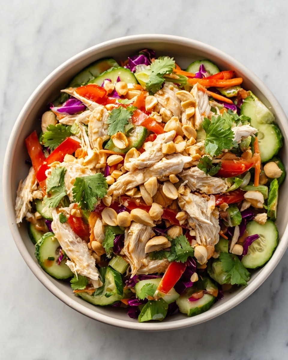 The dish is a vibrant, fresh salad served in a white bowl, filled with shredded white chicken pieces layered on top of a colorful mix of sliced cucumbers, thin carrot strips, red bell peppers, and purple cabbage. Scattered throughout are light brown peanuts and fresh green cilantro leaves adding pops of green. The textures vary from the crispness of the vegetables to the tender, juicy chicken, all combined to create a bright and appetizing look. The bowl sits on a white marbled surface. photo taken with an iphone --ar 4:5 --v 7