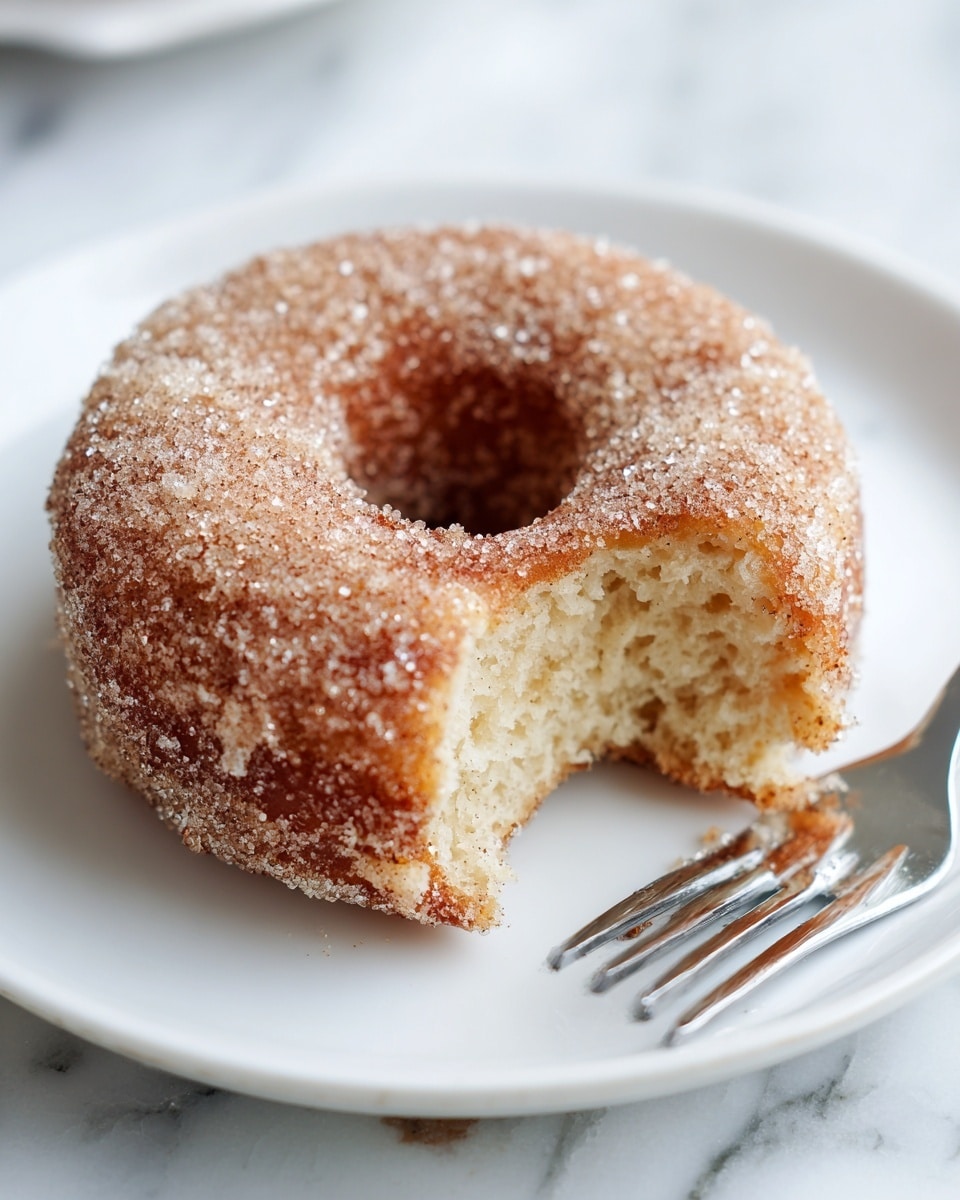 A close-up of a single round donut with a hole in the middle, coated in a thick layer of sparkling sugar crystals mixed with cinnamon, creating a textured, brownish-red sugary crust; one bite is taken out, showing the soft, fluffy, light beige interior with a slightly crumbly texture. The donut sits on a clean white plate with a shiny silver fork resting beside it on a white marbled surface. Photo taken with an iphone --ar 4:5 --v 7