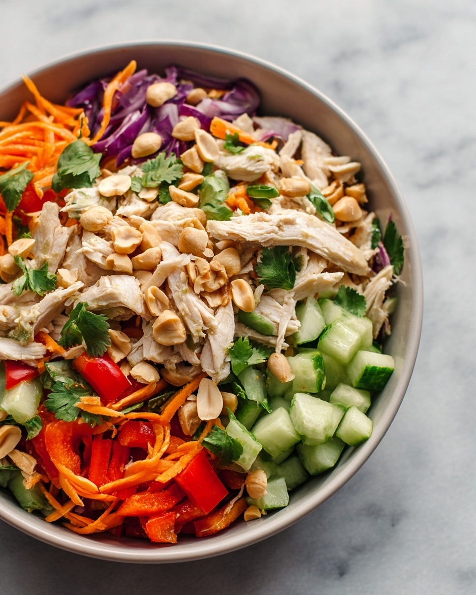 The image shows a close-up of a bowl filled with a colorful chicken salad. The bottom layer consists of chopped cucumber pieces that are light green with a smooth texture. On top of the cucumber, there are thin strips of bright orange carrots mixed with small bits of red bell pepper and thin slices of purple onion. The next layer features chunks of shredded white chicken spread evenly all over the salad. Scattered on top are halved toasted peanuts, adding a light brown crunchy texture. Small green cilantro leaves are spread throughout the dish, adding color and freshness. The bowl itself is white and set on a white marbled surface, creating a clean and fresh look. Photo taken with an iphone --ar 4:5 --v 7
