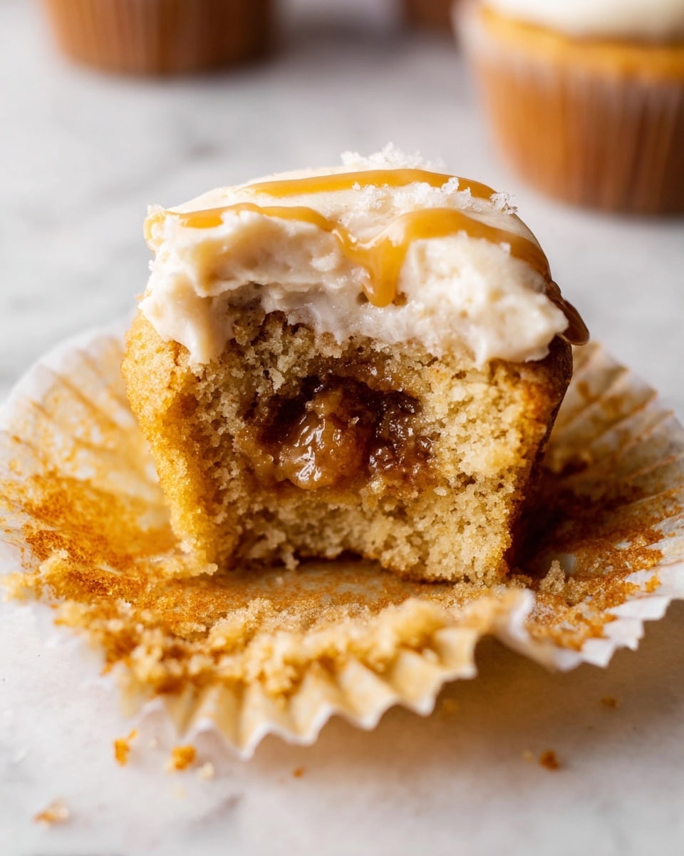 The image shows a close-up of a single cupcake with a light brown, soft texture that is split in half, revealing a fluffy inside topped with a dollop of smooth, white icing. These halves rest on a white cupcake liner with subtle folds, placed on a white marbled surface. Around the broken cupcake, there are whole cupcakes topped with the same smooth white icing, adding contrast with their round, precise shapes and uniform texture. The lighting is soft, showing the moist crumb and creamy frosting clearly. Photo taken with an iphone --ar 4:5 --v 7