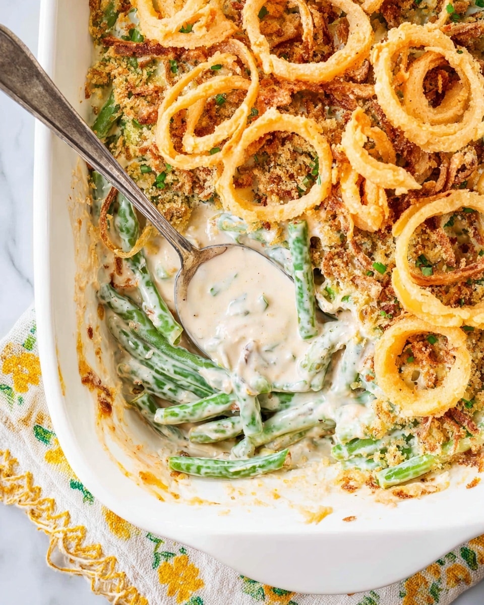 The dish is a creamy green bean casserole in a white ceramic baking dish with a white marbled texture underneath. The casserole has three main layers: the bottom layer is bright green green beans, the middle layer is a creamy, pale yellow sauce covering the green beans, and the top layer consists of golden brown, crispy fried onion rings scattered evenly with some orange breadcrumb bits. A silver spoon is lifting a portion, showing the green beans coated with sauce and topped with a few onion rings and breadcrumbs. The overall look is warm and inviting with a mix of green, creamy yellow, and crisp golden colors. Photo taken with an iphone --ar 4:5 --v 7