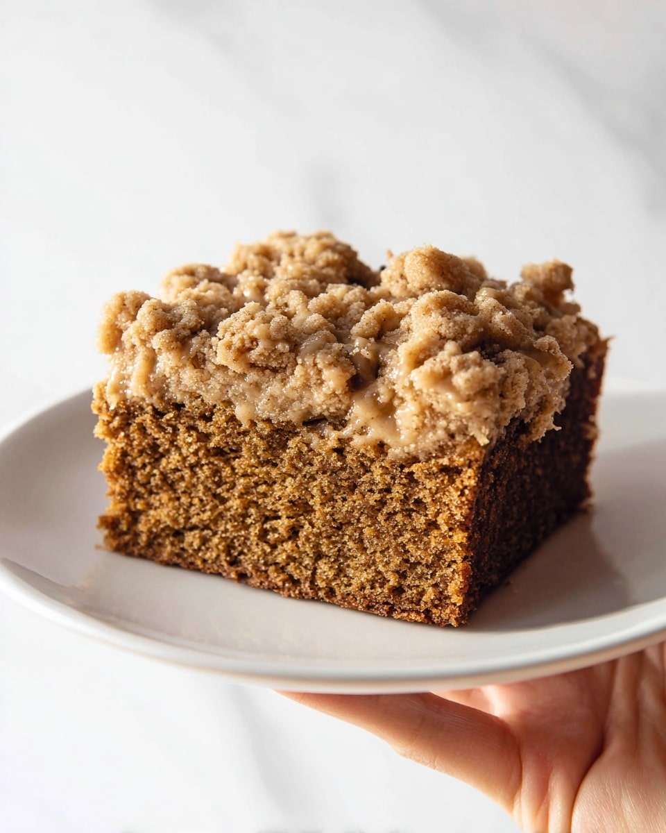 A thick square piece of cake is shown on a white plate, held by a woman's hand. The cake has two layers: the bottom layer is a dense, moist brown cake with a soft crumb texture, and the top layer is a lighter tan crumb topping with a streusel texture that looks crumbly and slightly glossy. The crumb topping has uneven chunks, adding a rough texture, and it slightly drips over the edges of the cake. The background and surface are a white marbled texture. photo taken with an iphone --ar 4:5 --v 7