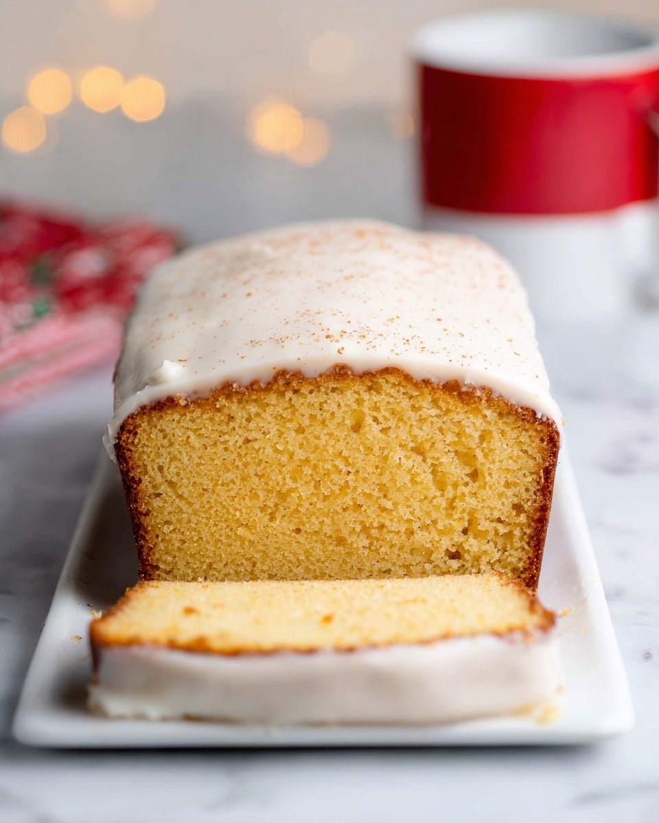 The image shows a loaf cake sliced to reveal its inside, placed on a white rectangular plate on a white marbled surface. The cake has two visible layers: the bottom layer is golden yellow, soft, and porous with a slightly darker brown crust on the sides, while the top layer is a thick, creamy white frosting that covers the entire top, dripping slightly down the sides. The frosting has a smooth texture with a light sprinkle of cinnamon or spice on top, adding tiny brown specks. The background has soft, blurred warm lights and a red and white mug that gives a cozy feeling. photo taken with an iphone --ar 4:5 --v 7