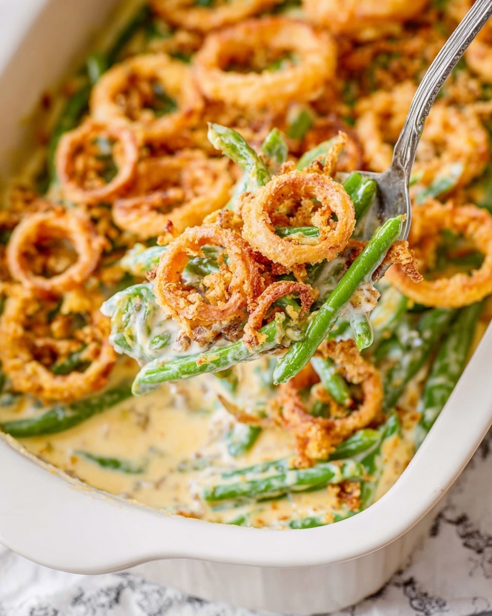 A white baking dish filled with a creamy green bean casserole. The bottom layer shows whole green beans covered in a smooth, white creamy sauce. On top of the sauce is a golden brown breadcrumb layer mixed with green herbs. The casserole is topped with large, crispy, light golden fried onion rings that are scattered unevenly. A silver spoon rests inside the dish, scooping out some of the creamy sauce and green beans, with some sauce splattered on the edges of the dish. The dish sits on a white marbled surface with a piece of fabric beside it featuring yellow floral patterns. Photo taken with an iphone --ar 4:5 --v 7