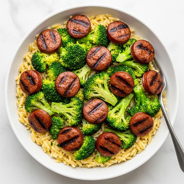 A white bowl filled with layers of small, light yellow orzo pasta at the bottom, topped with bright green broccoli florets that have a slightly crisp texture, and round, browned sausage slices scattered evenly on top showing char marks and a juicy inside, all placed on a white marbled surface with a fork visible in the bowl. Photo taken with an iphone --ar 4:5 --v 7
