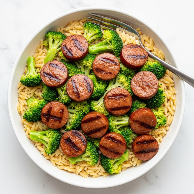 A white bowl filled with a dish that has three layers: the bottom layer is orzo pasta in a light yellow color and smooth texture, the middle layer has bright green broccoli florets, and the top layer shows golden-brown sliced sausage pieces with grill marks, slightly shiny, scattered evenly over the broccoli and pasta. The bowl sits on a white marbled surface, and a metal spoon is partially visible on the right side inside the bowl. Photo taken with an iphone --ar 4:5 --v 7