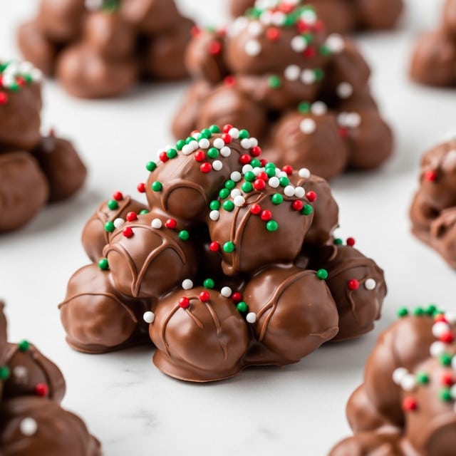 This image shows a cluster of milk chocolate coating on nuts or similar pieces, forming an uneven mound with several small and large lumps. The surface of the chocolate is shiny and smooth in some areas, with a glossy texture. Scattered over the top are tiny, round sprinkles in bright green, red, and white colors, adding a festive look. Other similar chocolate clusters are blurred in the background. All clusters are on a smooth white marbled surface. photo taken with an iphone --ar 4:5 --v 7
