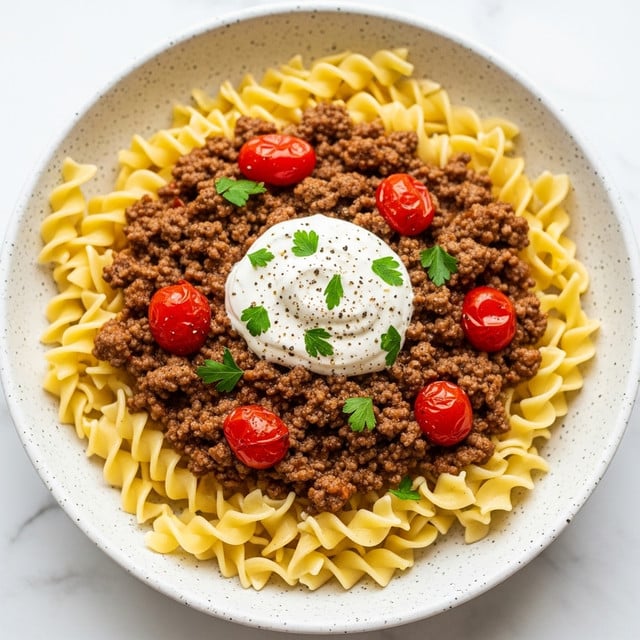 A white speckled bowl filled with several layers of food starts with a base of wavy, light yellow egg noodles. On top of this is a thick layer of browned ground beef mixed with small pieces of red cherry tomatoes and light brown sauce, spread evenly. At the center, a dollop of white sour cream is placed, sprinkled with small green parsley leaves and some black pepper. The dish sits on a white marbled surface. photo taken with an iphone --ar 4:5 --v 7