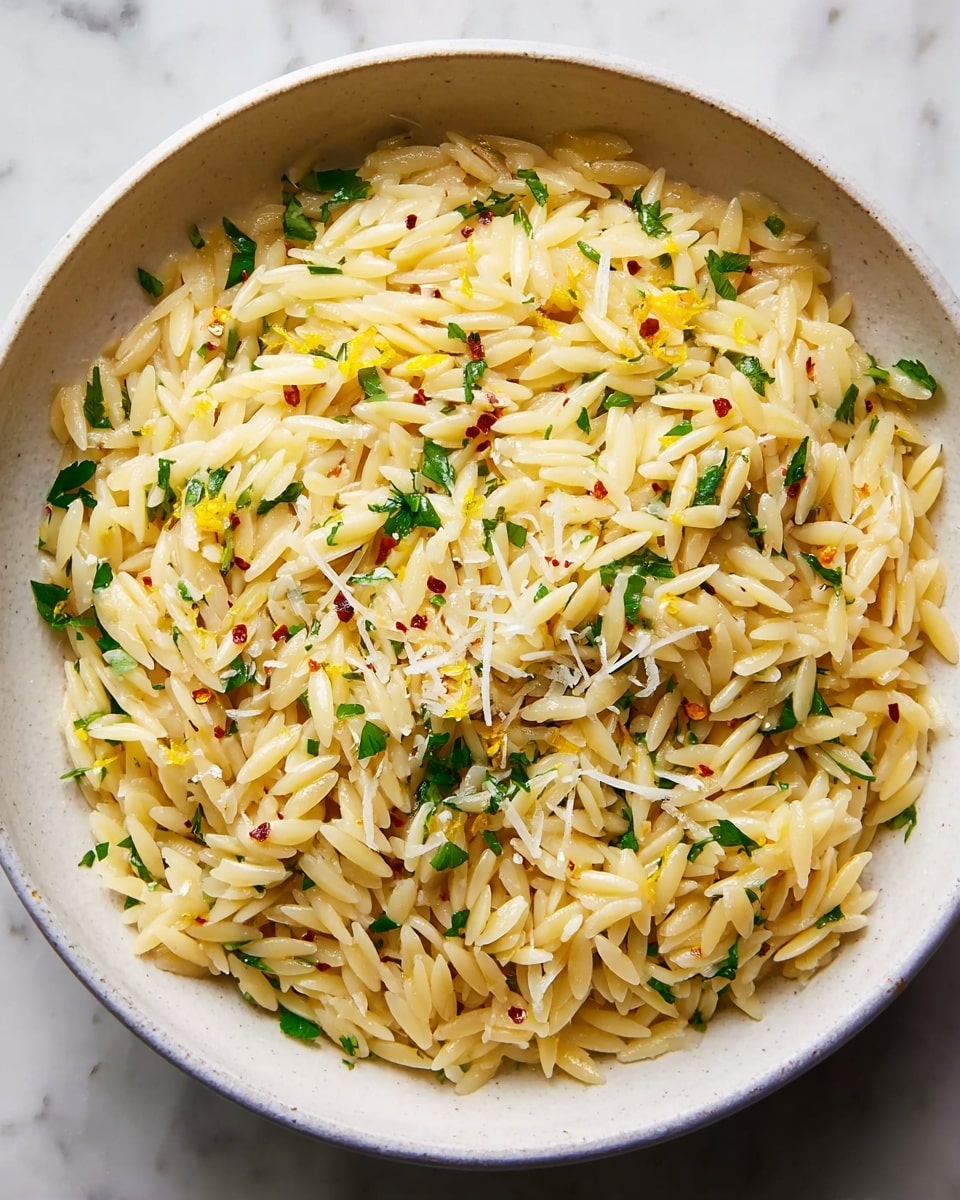 A white bowl filled with orzo pasta, cooked and mixed with finely chopped green herbs scattered evenly throughout. The pasta is light golden in color with a slightly glossy texture from oil or butter, and small bits of grated white cheese and yellow lemon zest are sprinkled on top. The surface is clean white marble, and the bowl is viewed from above, showing the uniform mix of ingredients. photo taken with an iphone --ar 4:5 --v 7