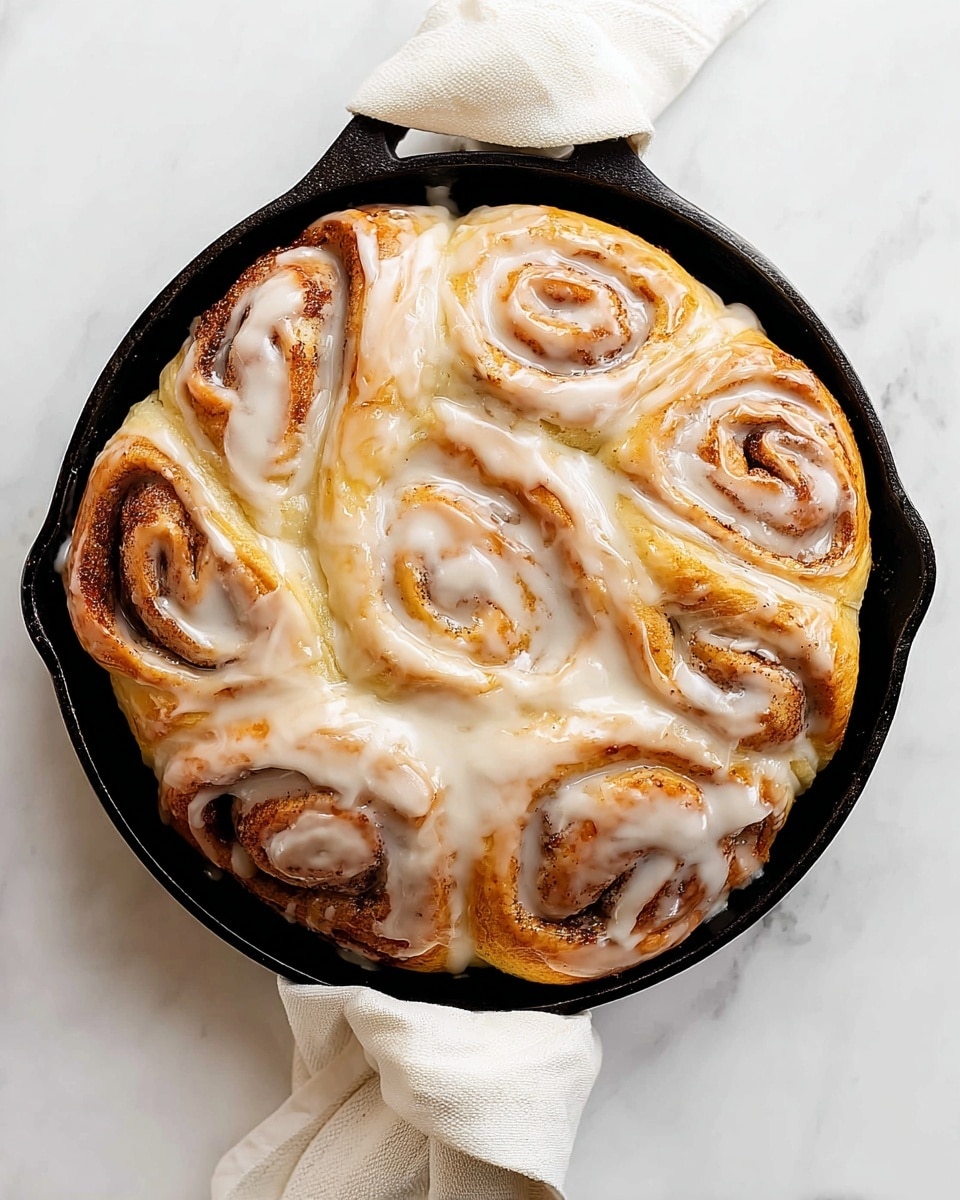 A large cinnamon roll is shown in a black cast iron skillet, placed on a white marbled surface with a white cloth wrapped around the handle. The roll has several thick, doughy layers spiraled inward, with a golden brown outer crust and light brown cinnamon filling visible between the layers. A shiny, creamy white glaze generously covers the entire roll, pooling slightly in the skillet. The glaze's smooth texture contrasts with the soft, fluffy dough beneath, making the roll look moist and rich. photo taken with an iphone --ar 4:5 --v 7
