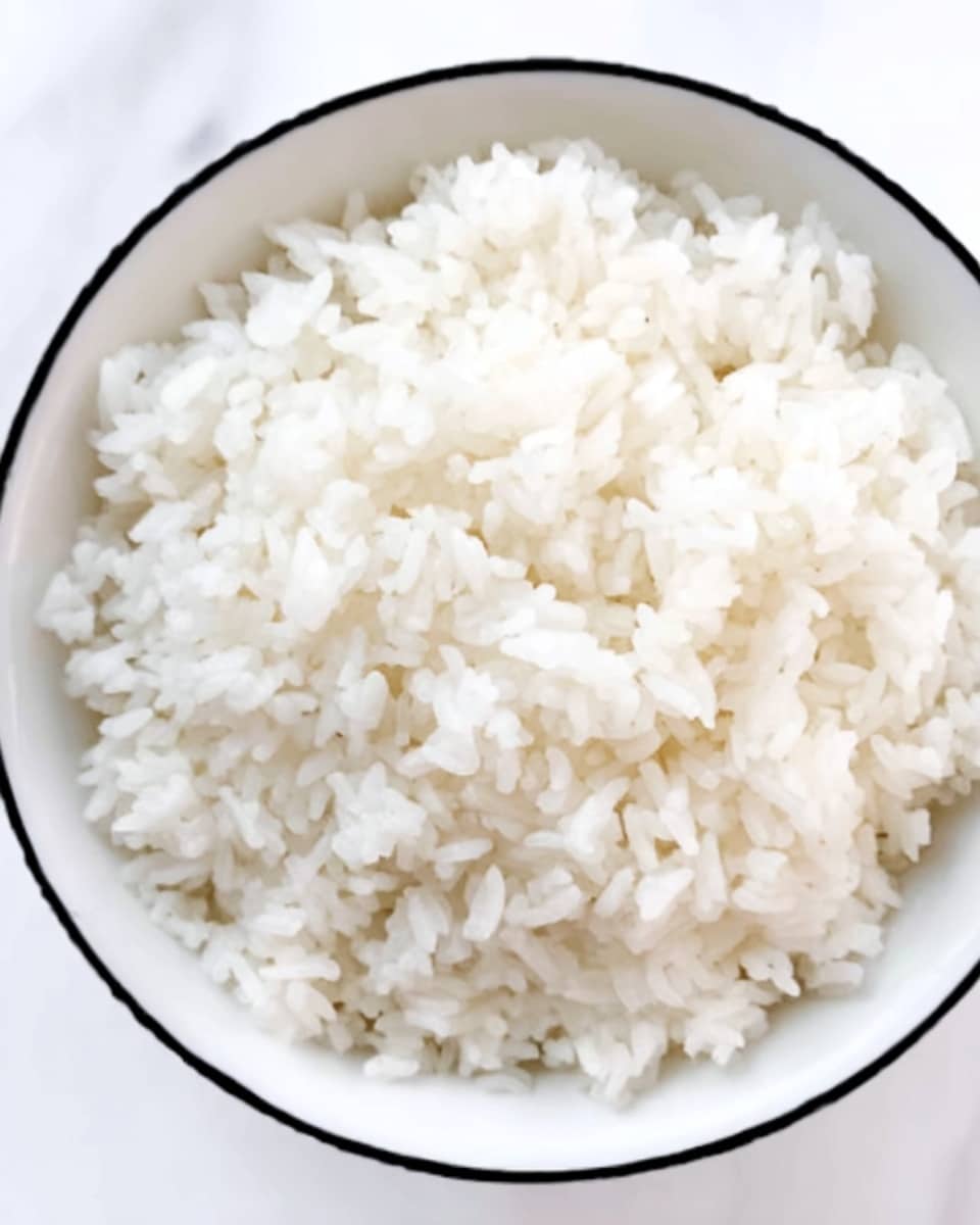 A close-up image of a white bowl filled with steamed white rice. The rice grains look soft, fluffy, and slightly sticky, filling the bowl almost to the top. The bowl has a thin black rim, and it sits on a white marbled surface, adding subtle texture in the background. The photo is simple, clear, and focused on the rice's texture and color with even lighting. Photo taken with an iphone --ar 4:5 --v 7