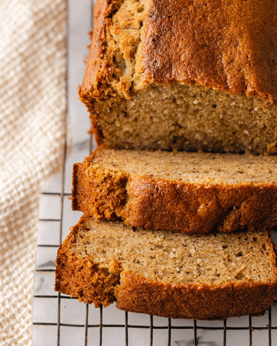 A loaf of banana bread is sliced into six thick pieces, stacked close together showing the golden brown, slightly cracked top crust and moist, light tan crumb inside. The texture looks soft and dense with small air holes throughout the bread's interior. The slices sit on a cooling rack atop a white marbled surface with a neutral, textured cloth just visible in the background. The warm tones of the bread contrast with the clean, light background. photo taken with an iphone --ar 4:5 --v 7