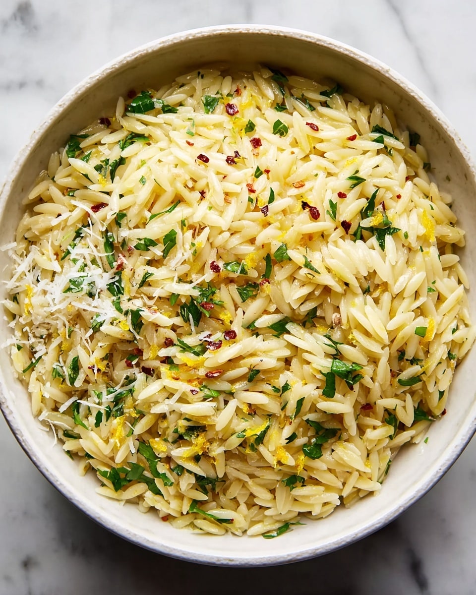 A white bowl filled with cooked orzo pasta, pale yellow in color with a slightly glossy texture, mixed with small pieces of green herbs scattered evenly throughout. On top of the orzo, there are fine shreds of white cheese and small specks of red pepper flakes with some bright yellow lemon zest sprinkled, adding color contrast. The bowl is set on a white marbled texture. photo taken with an iphone --ar 4:5 --v 7