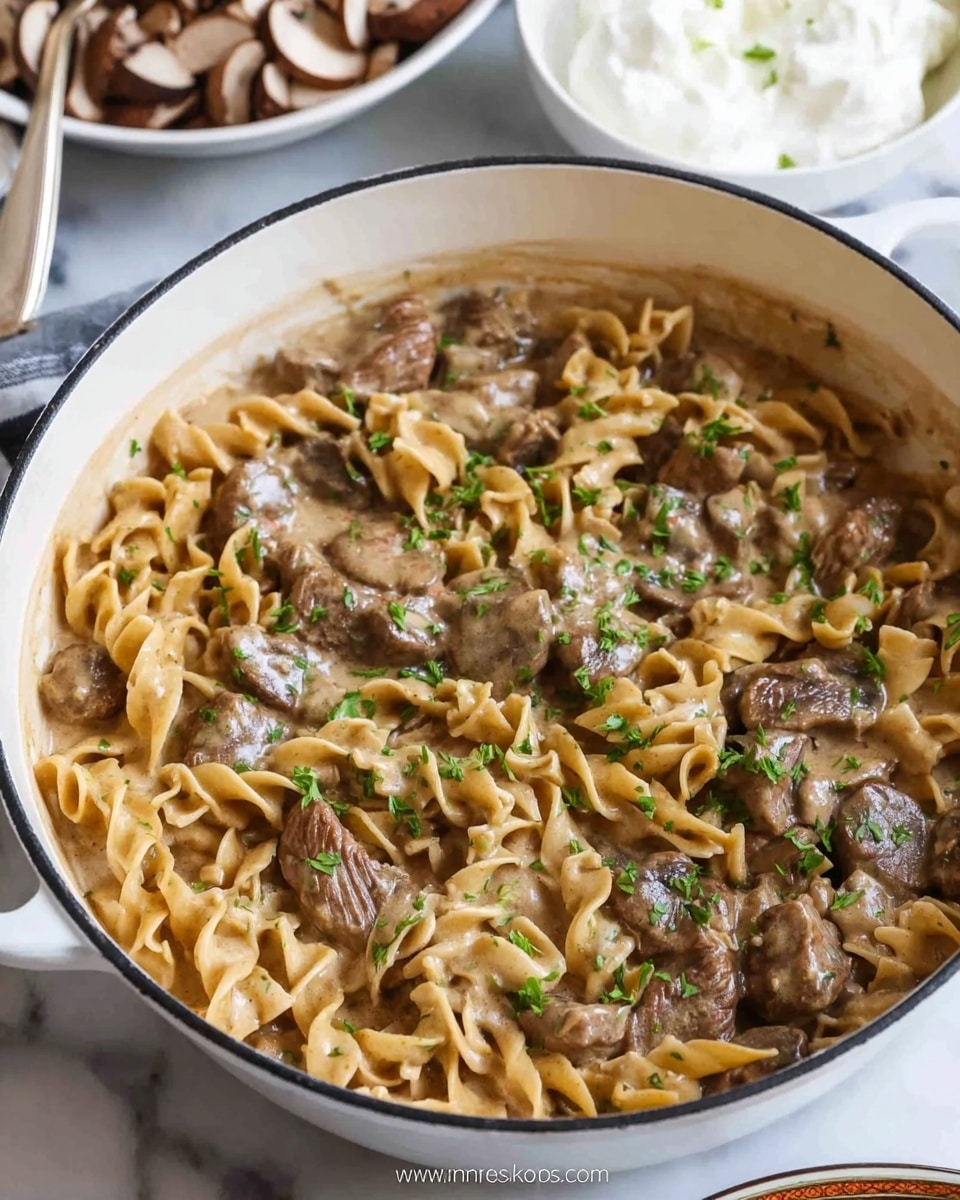 A close-up view of a white pot filled with a creamy beef stroganoff dish, showing two main layers: the bottom layer is light brown creamy sauce, and the top layer has twisted egg noodles mixed with pieces of browned beef and sliced mushrooms, all coated in the sauce; small green herb bits are sprinkled on top. The pot is placed on a white marbled surface, with a small white bowl of sour cream and a white bowl of sliced mushrooms in the background. photo taken with an iphone --ar 4:5 --v 7