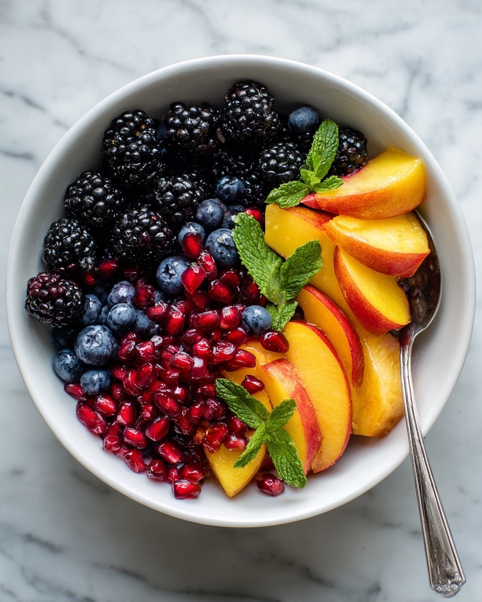 A white bowl filled with a fresh fruit salad showing three main layers: the bottom layer has dark blackberries with their bumpy texture, the middle layer is made up of bright yellow-orange peach slices with smooth skin, and the top layer is sprinkled with shiny red pomegranate seeds, small round blue blueberries, and thin green mint strips scattered over. A silver spoon is placed inside the bowl on the right side. The bowl sits on a white marbled surface. Photo taken with an iphone --ar 4:5 --v 7
