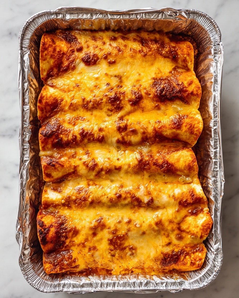 A close-up view of a foil tray filled with five baked enchiladas placed side by side, each covered with a thick layer of melted golden-brown cheese that looks slightly crispy on top. The cheese layer is bubbly with darker browned spots throughout, and a rich reddish-brown sauce can be seen peeking through cracks in the cheese. The foil edges have crinkled texture, and the tray sits on a white marbled surface. photo taken with an iphone --ar 4:5 --v 7