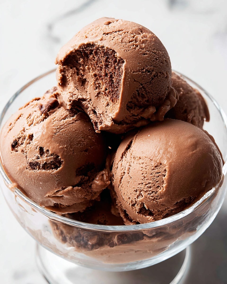 A glass bowl filled with four scoops of rich chocolate ice cream, each scoop showing a creamy, smooth texture with some soft fluffiness and small air pockets. The scoops are stacked unevenly, with one scoop resting partly on top of another, showcasing different shades of dark brown. The bowl is clear, letting the chocolate ice cream's color and texture be seen through its curved edges. The background features a soft white marbled texture, adding a clean and simple look. photo taken with an iphone --ar 4:5 --v 7
