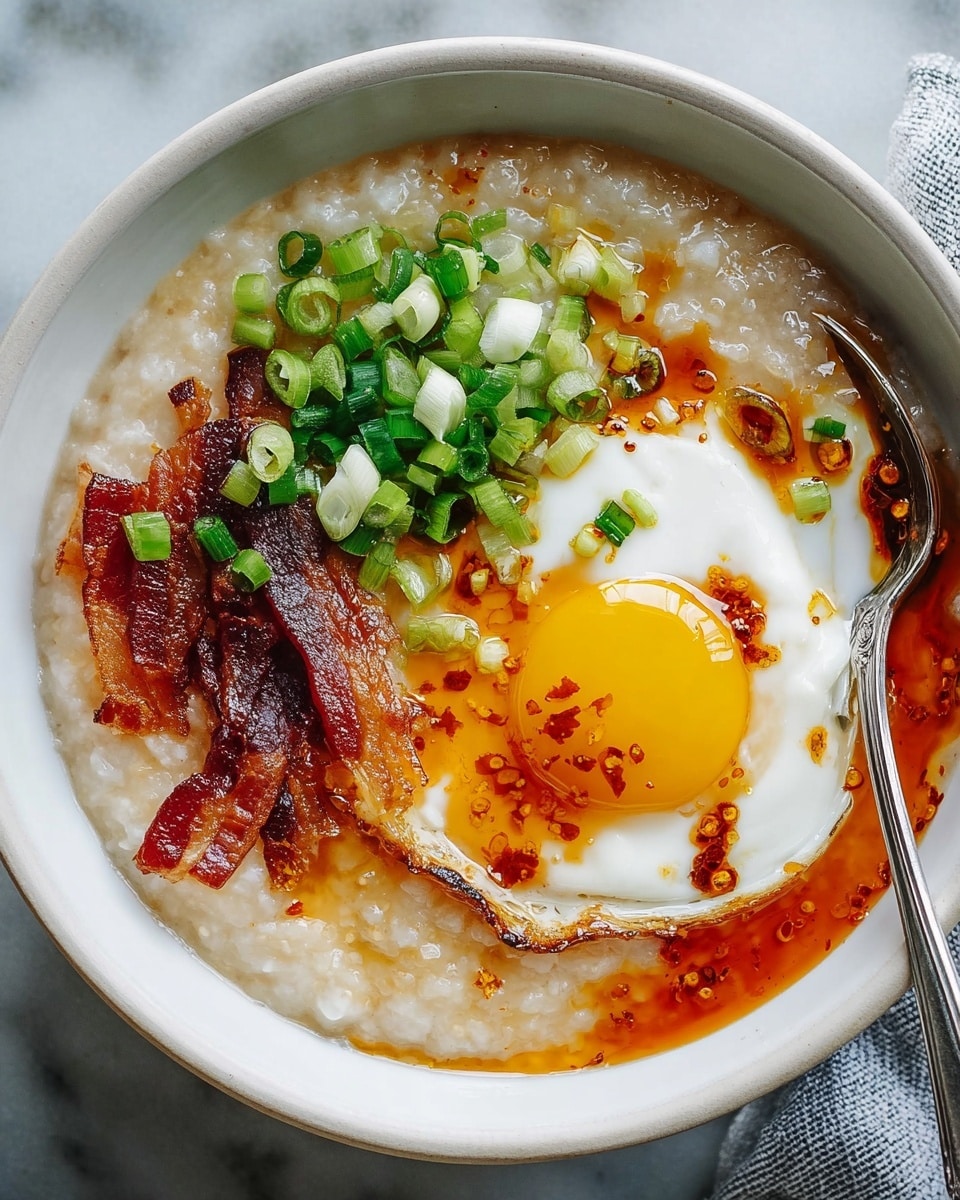 A white bowl filled with a thick, light beige rice porridge as the base layer, topped on one side with chopped bright green scallions, next to dark brown, crispy strips of bacon, and a sunny-side-up egg with a bright yellow runny yolk and a white cooked edge. A reddish-brown chili oil sauce is drizzled partly over the egg and some bacon, adding a shiny texture and vibrant color contrast. A metal spoon rests inside the bowl, and the bowl is placed on a white marbled surface. Photo taken with an iphone --ar 4:5 --v 7