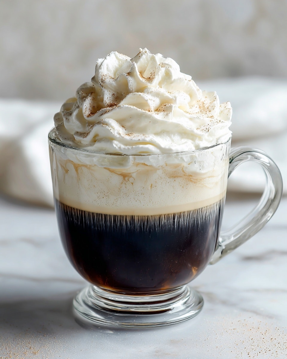 A clear glass cup filled with two layers: the bottom layer is a smooth light brown coffee, and the top layer is a thick white foam layer. On top of the foam, there is a large swirl of white whipped cream sprinkled with light brown cocoa powder. The glass cup sits on a white marbled surface, with a blurred dark background behind it. Photo taken with an iphone --ar 4:5 --v 7