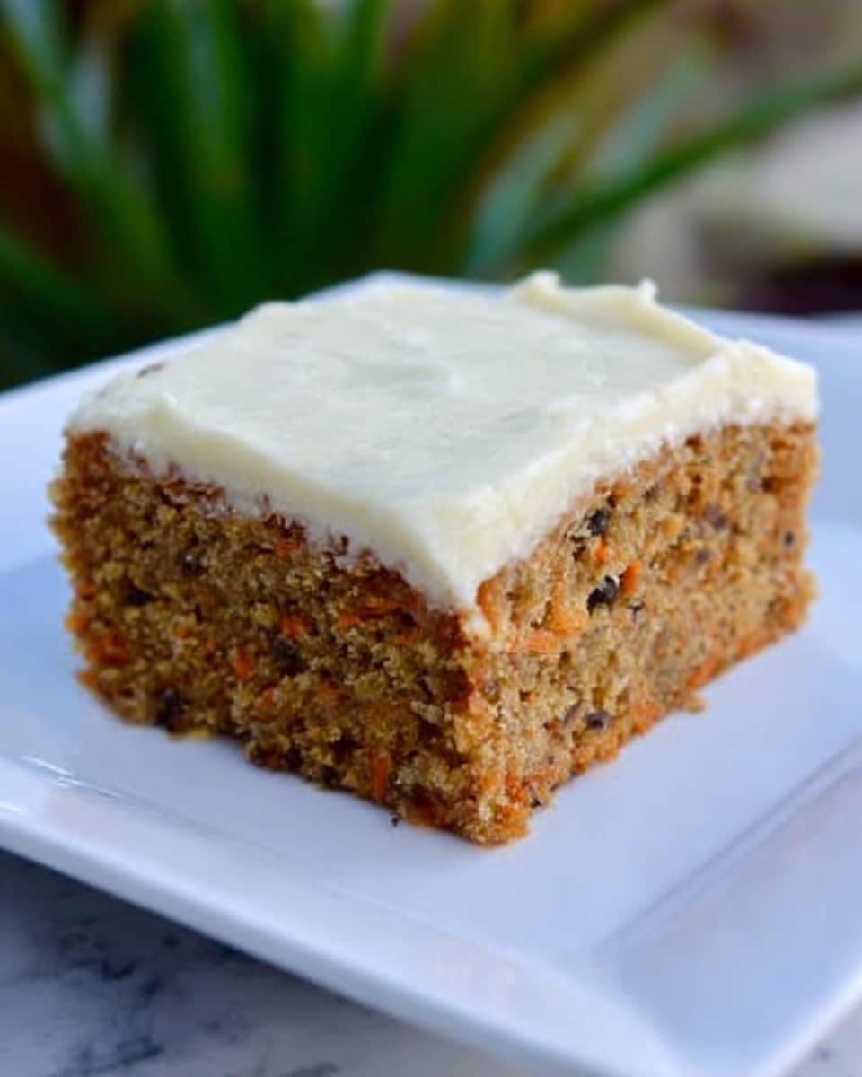 A single square piece of moist-looking carrot cake with visible small bits of carrot and dark spots of raisins or nuts is sitting on a white plate. The carrot cake has a light brown color and is topped with a smooth, thick layer of creamy white frosting. The texture of the frosting looks soft and slightly shiny. The background shows a white marbled surface with some greenery blurred in the distance. photo taken with an iphone --ar 4:5 --v 7