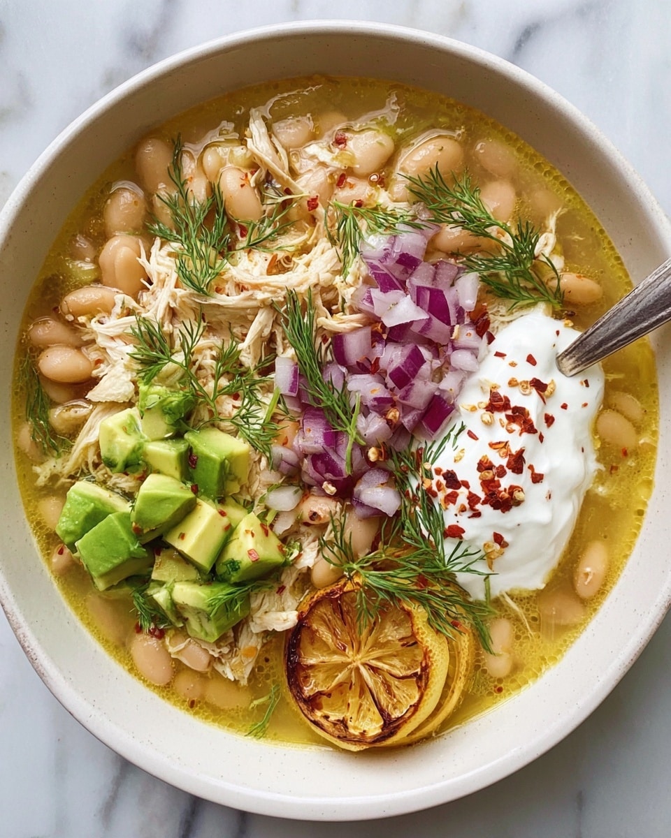 A bowl filled with a light yellow broth as the base layer, with visible beans and shredded chicken mixed inside. On top, there are diced purple onions scattered in the center, fresh green dill sprigs placed around, and chunks of bright green avocado pieces on one side. A dollop of white sour cream sits beside the avocado with sprinkled red chili flakes over it. Two lemon wedges with a roasted texture rest near the sour cream, adding a warm yellow tone. A spoon is placed at the edge of the white bowl, which sits on a white marbled surface. photo taken with an iphone --ar 4:5 --v 7