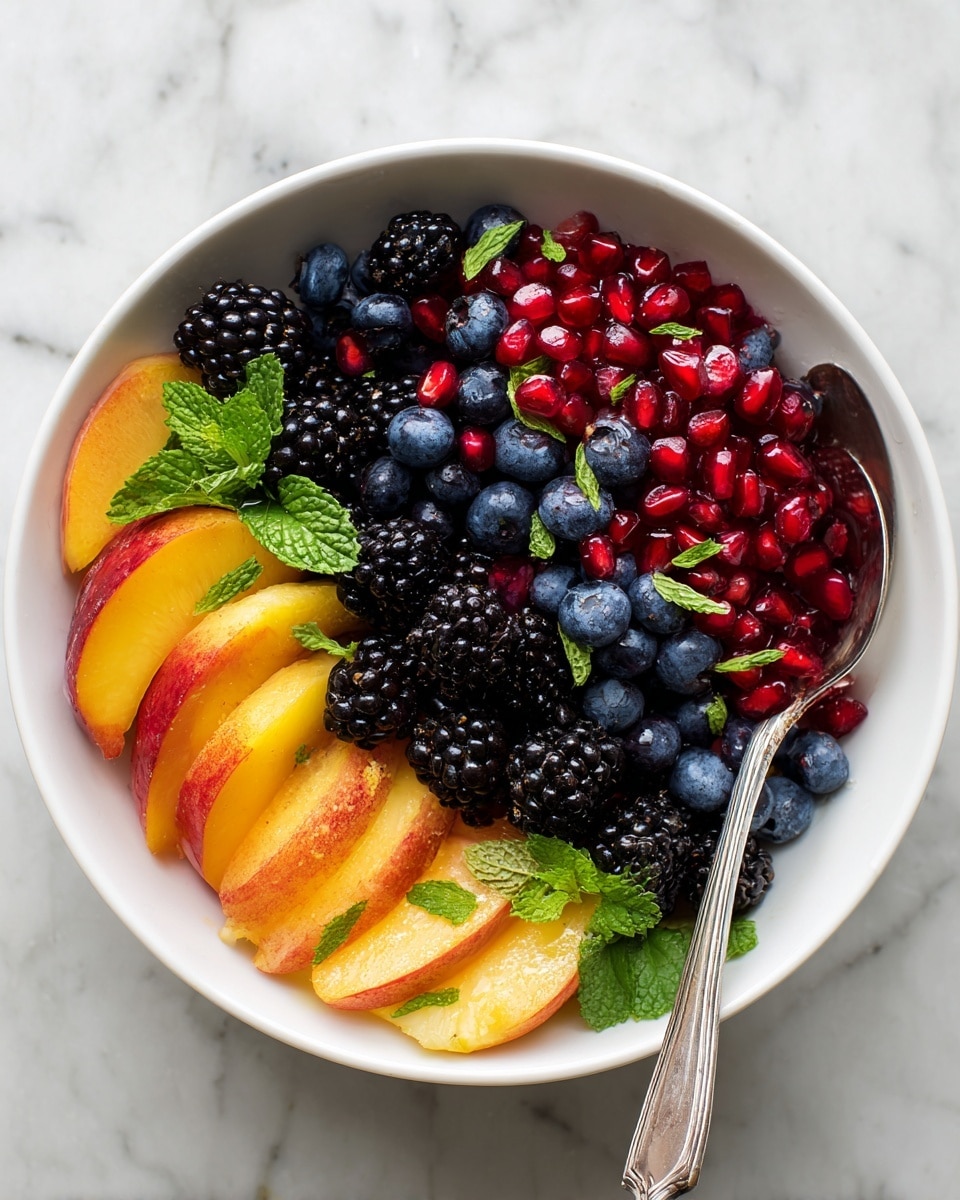 A white bowl filled with a colorful fruit salad showing three main layers: the base layer has yellow-orange peach slices with a smooth, slightly shiny texture arranged around the bowl's edge and throughout; the middle layer is made of deep purple-black blackberries, plump and textured, mixed evenly with small, round, dark blue blueberries; on top, bright red pomegranate seeds add a glossy, jewel-like detail sprinkled all over with thin, fresh green mint strips scattered lightly for contrast. The bowl sits on a white marbled surface, and a silver spoon peeks from the right side. photo taken with an iphone --ar 4:5 --v 7