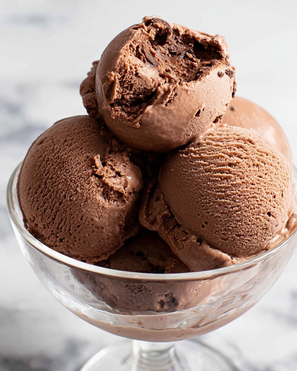 Four scoops of rich, creamy chocolate ice cream are placed inside a clear glass bowl. The scoops show a smooth, dense texture with visible freezing crystals and soft swirls, giving them a fresh and inviting look. The top scoop has a slightly bitten side revealing the soft and smooth inside. The bowl rests on a surface with a white marbled texture, enhancing the contrast with the dark brown of the ice cream. photo taken with an iphone --ar 4:5 --v 7